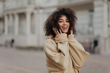 Brunette curly dark-skinned woman listens to music in headphones outside. Attractive lady in beige hoodie walks outdoors. - obrazy, fototapety, plakaty