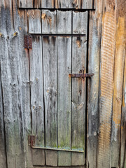 A wall with a door on rusty rickety iron hinges of an old wooden barn in the village, the door is closed on a rusty latch, the boards at the top of the door have deteriorated over time background