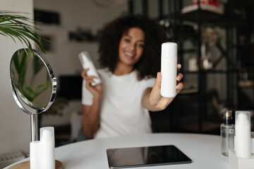Happy dark-skinned woman holds two bottles of skin care creams. Close-up photo of white face lotion. Blurred effect.