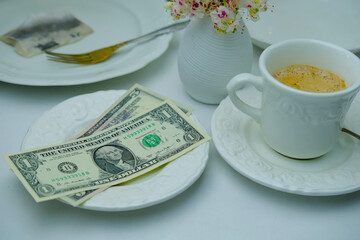 close-up of white plate for money, American dollars banknotes and coins, Restaurant bill, cup of coffee, delicate pink flowers, dishes with food, the concept of tip money, change of the waiter