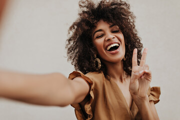 Cheerful woman shows peace sign on grey background. Dark-skinned curly lady in brown blouse takes...