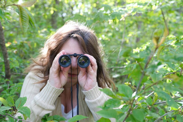 cunning curious European adult woman close-up, holding black field binoculars with zoom, hiding in...