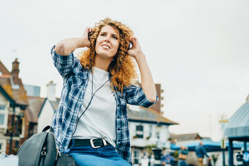 Naklejka premium Happy caucasian woman with red curly hair in headphones listening to music on a city street. Bottom view. Selective focus, copy space