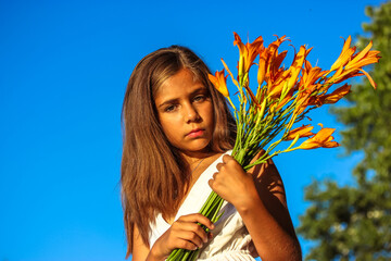 A beautiful serious teenage girl in white dress with a bouquet of orange lilies against a blue sky...