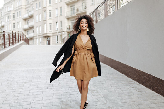 Beautiful dark-skinned woman walks outside. Curly brunette lady in black coat and brown dress moves outdoors.