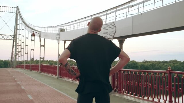 Young white Man in black sport uniform doing warm-up neck before run pedestrian bridge at dawn. High quality 4k footage