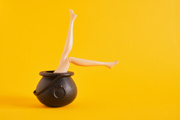 doll legs sticking out of small plastic witch cauldron on yellow background, halloween concept