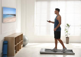 Fototapeta na wymiar Sporty man with remote control training on walking treadmill and watching TV at home