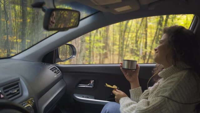 Brunette woman in warm sweater hold yellow leaf and drinking tea inside car while enjoying picturesque view autumn forest out of window. Travelling by car concept