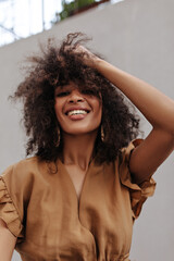 Fototapeta na wymiar Brown-eyed curly woman in brown dress ruffles hair. Stylish dark-skinned lady in beige blouse smiles and poses outside.
