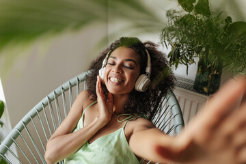 Good-humored dark-skinned woman in silk green dress listens to music in headphones, takes selfie and sits on chair at home.