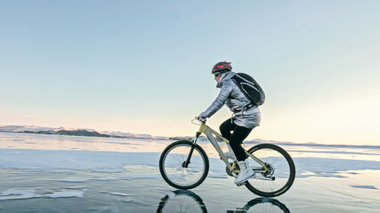 Fototapeta premium Woman is riding bicycle on the ice. Girl is dressed in a silvery down jacket, cycling backpack and helmet. Ice of the frozen Lake Baikal. Tires on bike are covered with spikes. Traveler is ride cycle.