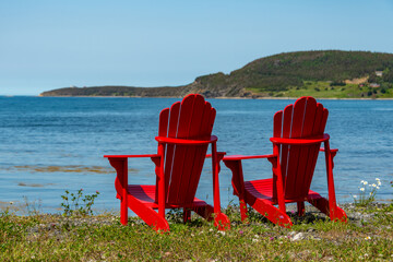 Two bright empty red Adirondack chairs on the edge of a green grassy meadow overlooking the blue ocean with mountains covered in trees, a bay, and a cove. The water is calm and the sky is blue. 