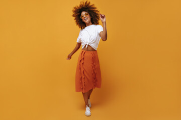 Modern girl in orange skirt and white t-shirt posing on isolated background. Happy lady in glasses...
