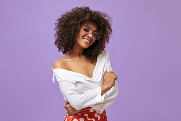 Cute lady with fluffy hairstyle in purple glasses poses on isolated backdrop. Optimistic girl in blouse and red pants smiles on lilac background..