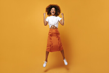 Cheerful girl with curly hair in orange skirt jumping on isolated backdrop. Charming woman in light t-shirt smiling on yellow background..