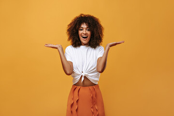 Positive girl in summer outfit smiling on isolated backdrop. Woman in white t-shirt and orange skirt pointing to place for text on yellow background..