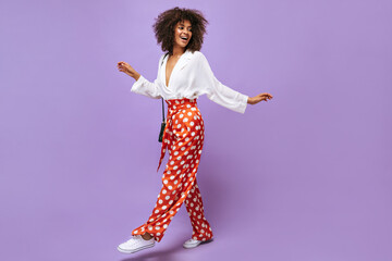Stylish lady in wide pants, sneakers and blouse posing on isolated backdrop. Trendy woman with wavy hair smiling on lilac background.. - Powered by Adobe