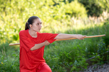 beautiful teen girl in red sports wushu uniform is training kung fu in the park, fighter junior...
