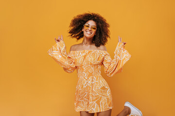 Positive lady in sunglasses smiles and jumps on isolated background. Wavy haired girl in jumpsuit showing peace signs on yellow backdrop..