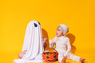 Children in mamia and ghost costumes for halloween sit in front of basket of sweets on yellow...