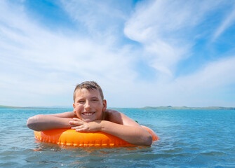 Boy swim in a blue lake at summer sunny day. He floats with orange floating ring and smile.