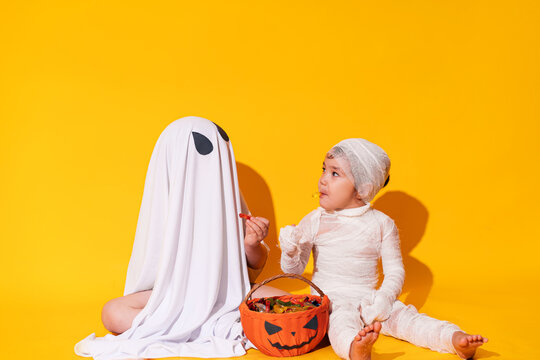 Children in mamia and ghost costumes for halloween sit in front of basket of sweets on yellow background