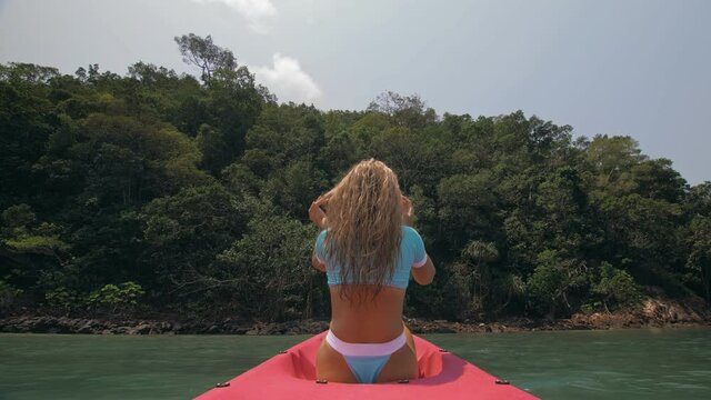 Sexy young woman in blue swimsuit swings feet in water on pink canoe on sea against hills backside view. Traveling to tropical countries. Attractive sportive girl is sailing on kayak in ocean.