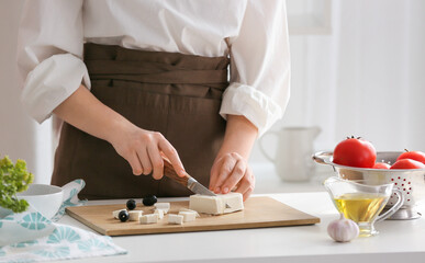 Woman cutting tasty feta cheese on table in kitchen, closeup