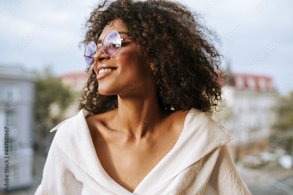 Wall mural beautiful woman with wavy hair in light clothes smiling outside. brunette curly girl in round eyegla