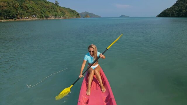 Pretty sportive blonde lady in stylish swimsuit sails on pink plastic canoe along azure ocean bay at exotic resort. Traveling to tropical countries. Girl is sailing on kayak in ocean, view from above.