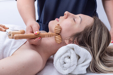 The hands of the masseur massages the face and neck of the girl using a wooden roller spiked...
