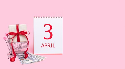 A gift box in a shopping trolley, dollars and a calendar with the date of 3 april on a pink...