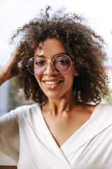 African woman with short wavy hair in lilac glasses smiling on balcony. Cute girl in white shirt looks into camera on terrace..