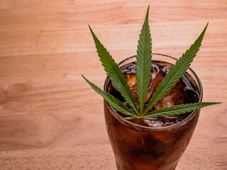 Glass of cola with ice cubes and cannabis leaf on the wooden table.