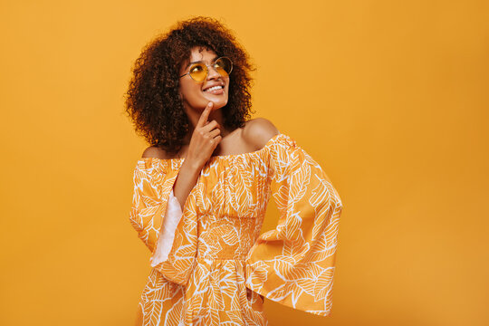 Dark-skinned woman in sunglasses smiling on isolated backdrop. Pensive girl in summer clothes posing on yellow background..
