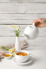 Woman pouring tasty chamomile tea from teapot into cup on light wooden background