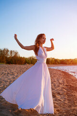 Fototapeta na wymiar beautiful slender young woman of Caucasian ethnicity with long hair stands in a white dress in the wind near the sea at sunset