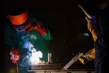 a factory worker wearing a green mechanic coveralls and safety helmet welding metalwork at night...