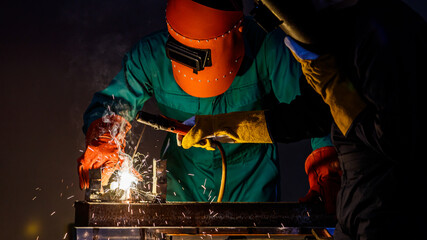 a factory worker wearing a green mechanic coveralls and safety helmet welding metalwork at night time in a factory while his colleague looking at a spark from welding