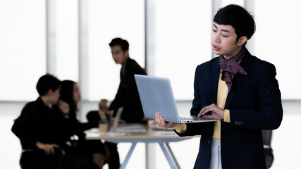 Portrait of LGBTQ transgender man office employee in casual suit standing using labtop notebook computer with elegant . and happy gesture with team colleagues blur in background
