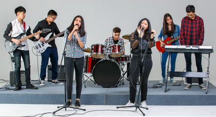 Group shot of teenage musicians playing the music and singing together on the weekends. Junior students play electric guitar, drum, and keyboard in the studio with the main vocalist singing in front