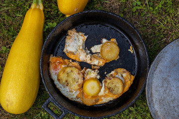 an old black round cast-iron frying pan. Yellow zucchini in round pieces with fried eggs. There is a large yellow zucchini nearby. On the background of green grass