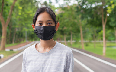 Portrait Asian pretty teen girl in casual dress wears black protective mask standing on footpath in public park under sunlight during coronavirus epidemic. Healthy lifestyle. Pollution free. - 447825837