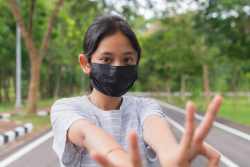 Portrait Asian pretty teen girl in casual dress wears black protective mask posing gesturing with her fingers while standing on the road in public park during COVID-19 epidemic. Healthy lifestyle.  - 447825866