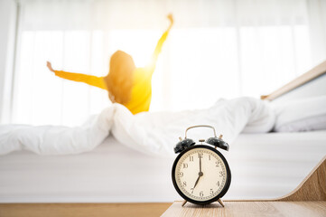 Good morning new day. Alarm clock wake up and woman sitting body stretch on bed beside window in...