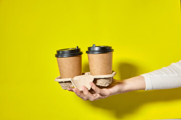 Hands holding two cups brown paper with black lid. Two coffee special offer or promo. Hands holding two cups on yellow screen background. Tea or Coffee to go. Brown paper cup with black lid.