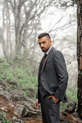attractive man with beard in business suit stands at tree in forest.