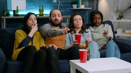 Multi-ethnic friends relaxing on sofa late at night during home party changing channel on television using remote while drinking beer. Group of multiracial people enjoying time together