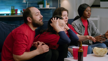 Support football cheering together while watching soccer on television. Multiethnic friends dissapointed after team losing match championship while sitting on sofa late at night in living room